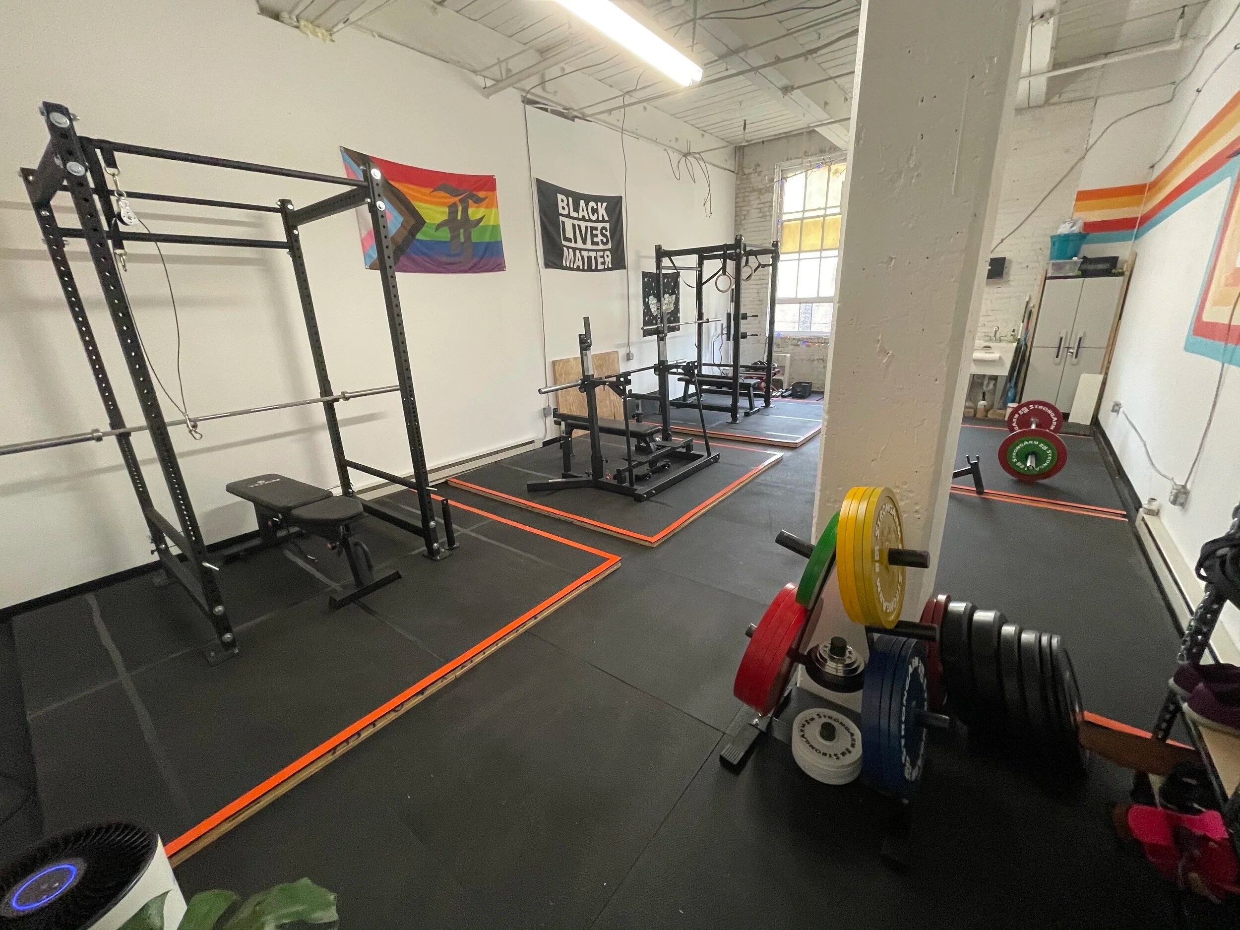 A gym with squat racks, bench press, weight plates, and LGBT and Black Lives Matter flags.