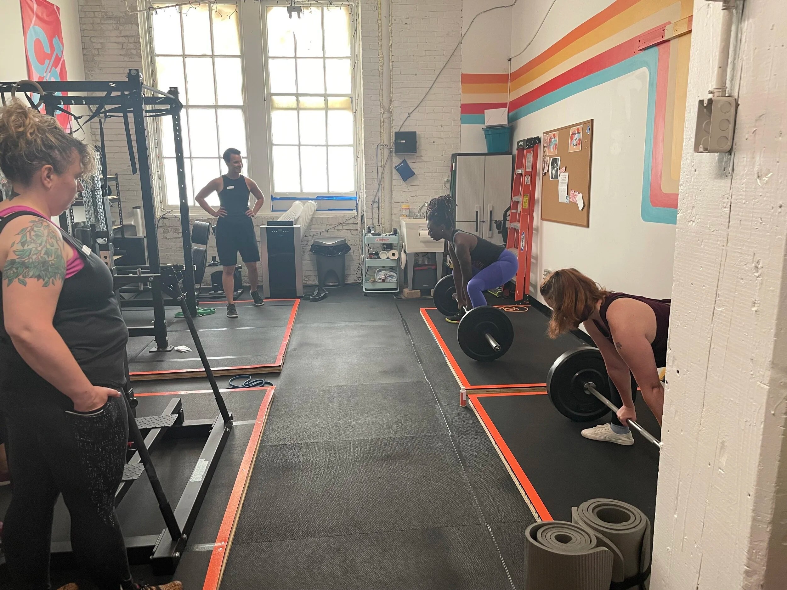 People in a gym setting, with two individuals preparing to lift barbells, observed by others.