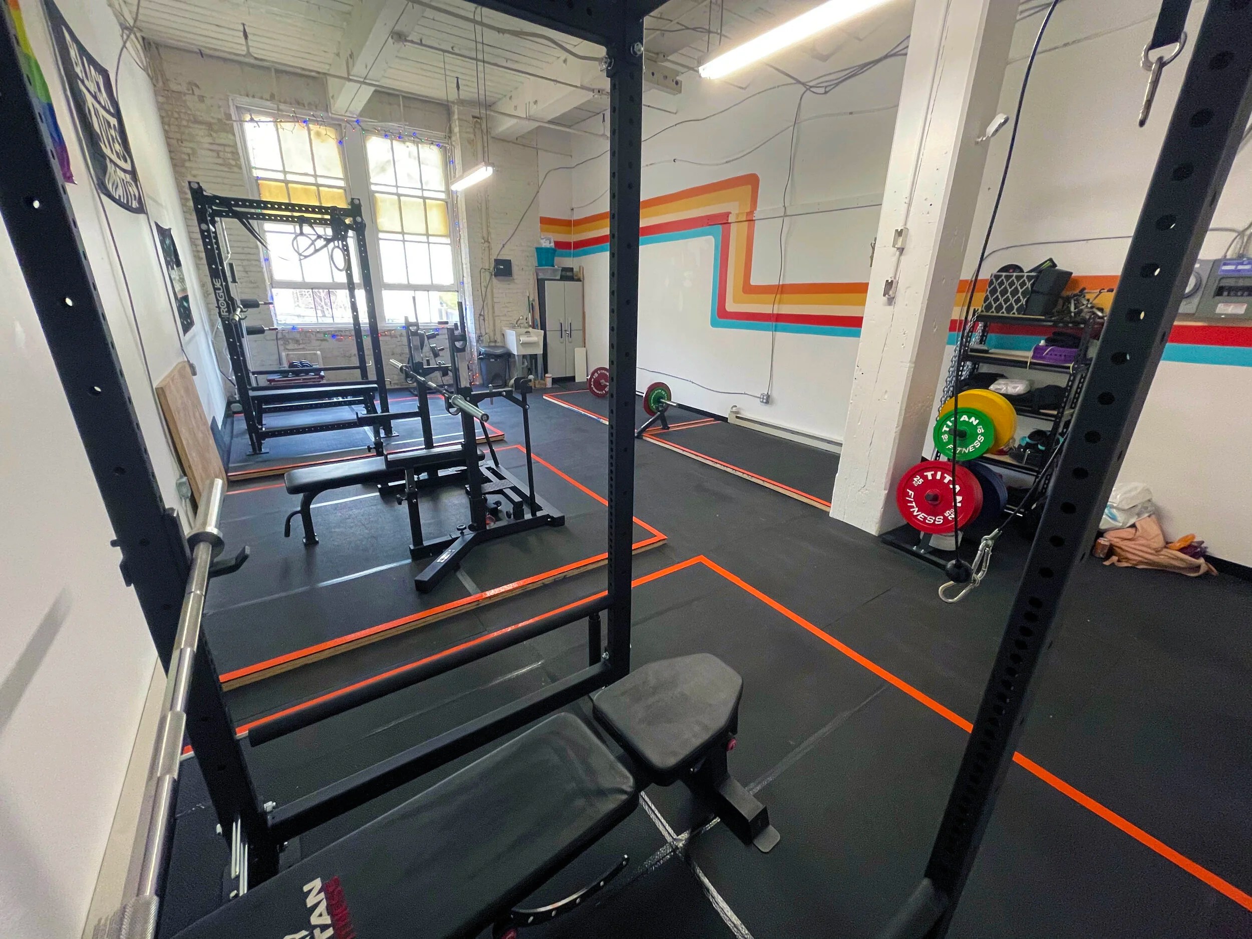 Interior of a gym with weightlifting equipment, including benches, squat racks, and colored weight plates. Walls feature colorful stripes and large windows are visible.