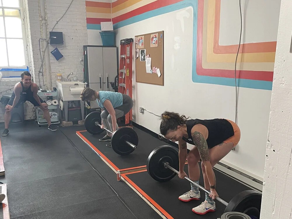Two women preparing to lift barbells in a gym with a trainer observing.